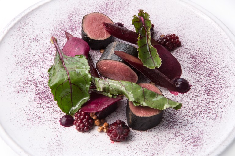 Plated circular cuts of smoked roe deer, surrounded by red berries and topped with green leaves, all sat on a red plate with red markings