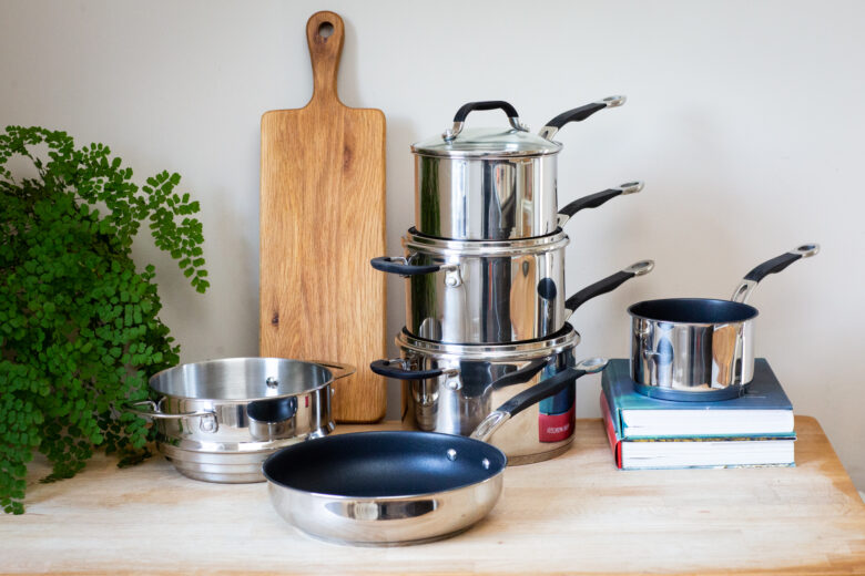 A set of our Stainless Steel Base set of pans, including a steamer and non stick saute pan in the foreground, and three saucepans stacked one on top of the other, next to a chopping board in the background.